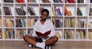 brown-skinned Black woman sitting crossed-leg in front of a bookshelf reading
