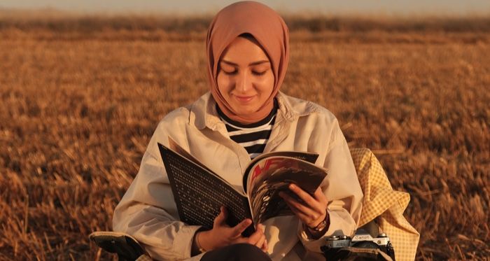 fair-skinned woman wearing a pink hijab sitting and reading in a hay field