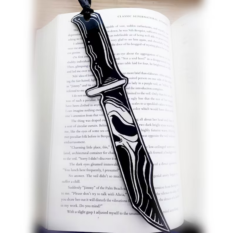 Photo of an open book with a bookmark placed on top of the pages. The bookmark is shaped like a knife, and has the famous Ghostface mask drawn on it in black and white. 