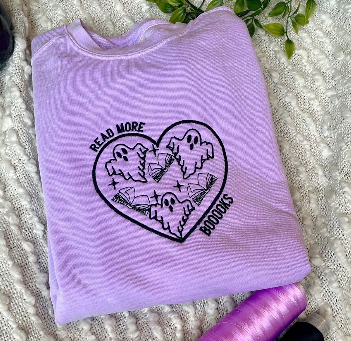 a purple sweatshirt with black embroidery. There is a heart with ghosts and books inside and text outside the heart says "read more booooks."
