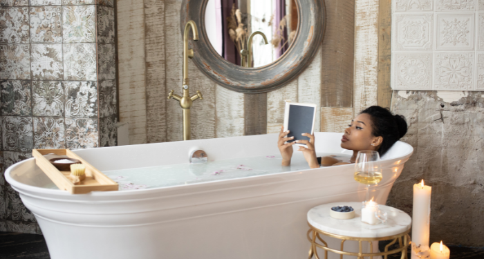 a photo of a Black woman reading in a bath with tapered candles burning and a glass of wine and bowl of blueberries beside the bathtub