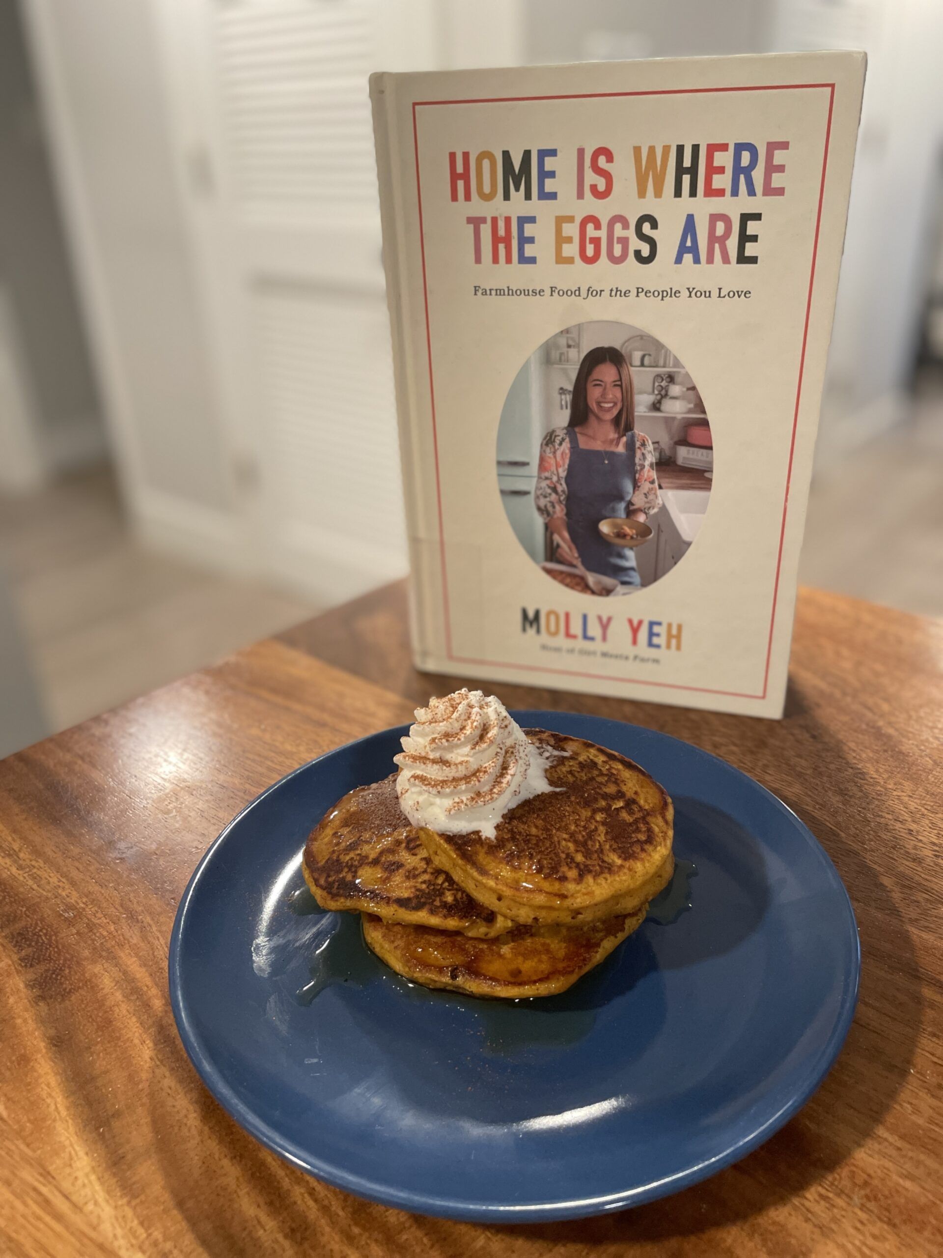 A small blue plate of orangey brown pancakes topped with syrup and whipped cream on a wooden table by the cookbook Home is Where the Eggs Are