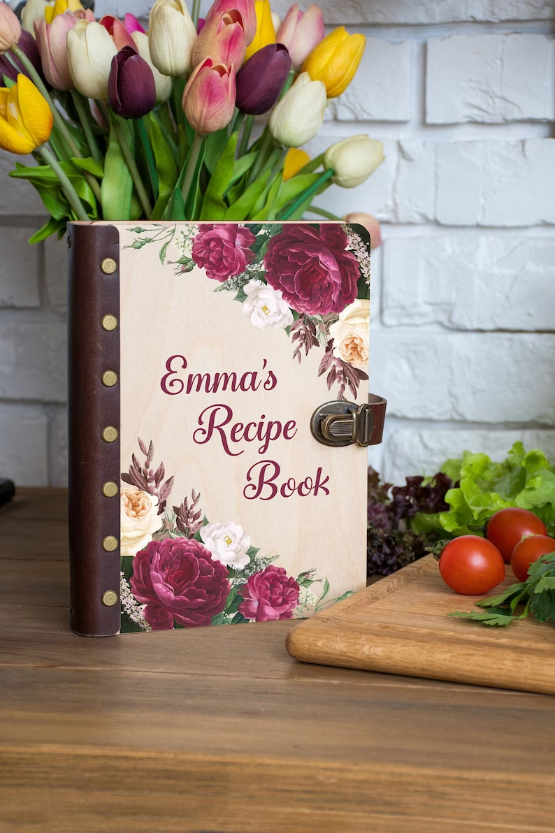 Photo of a recipe notebook with a wooden cover and a leather spine and claps standing in front of a vase of flowers, the cover has flowers at the corners and text in the middle