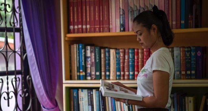 a tanned-skin Asian woman standing in front of a bookshelf reading a book