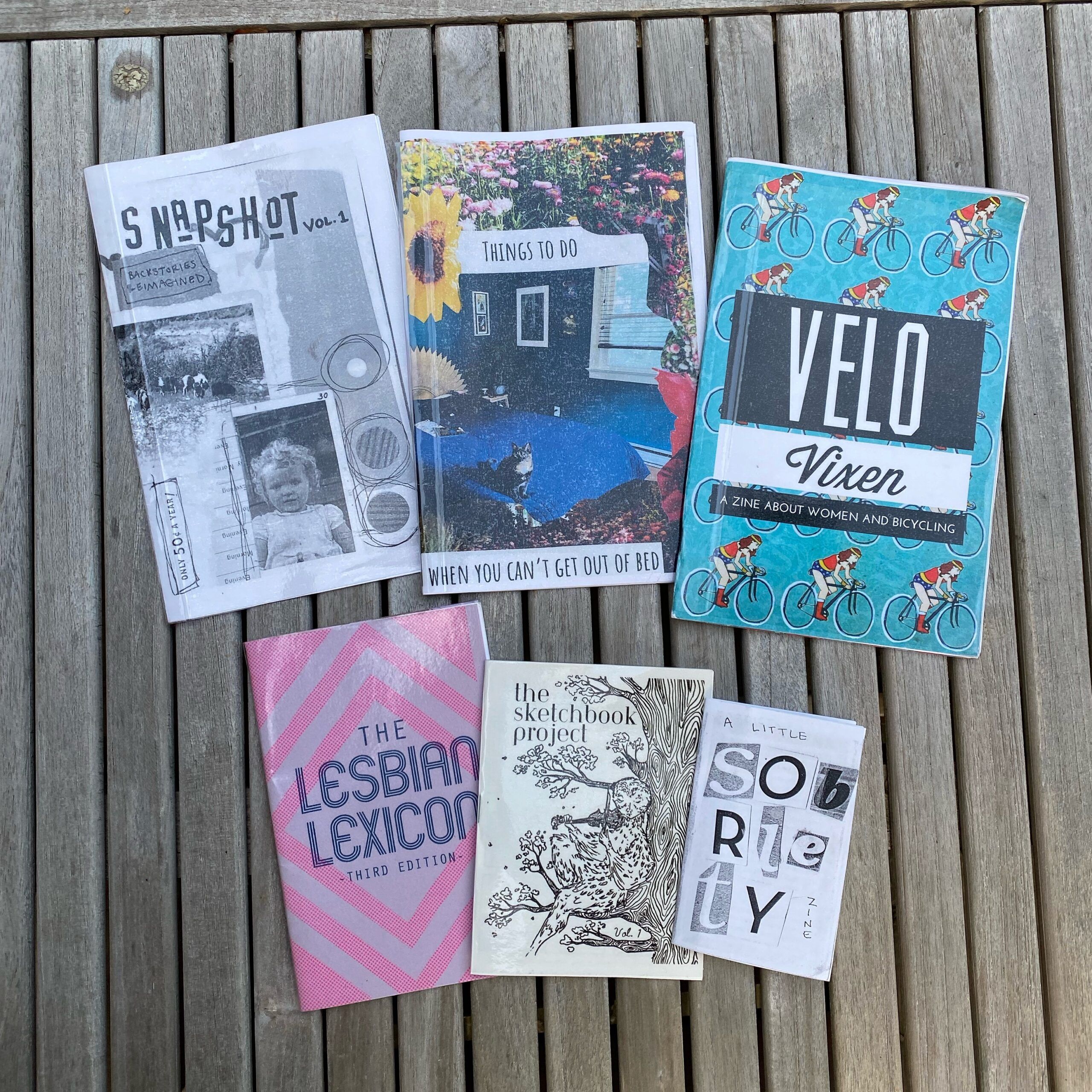 Six zines of different sizes and colors spread out on an outdoor table.