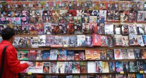 A person with short dark hair, wearing a red coat, stands in front of a wall displaying many different comic books