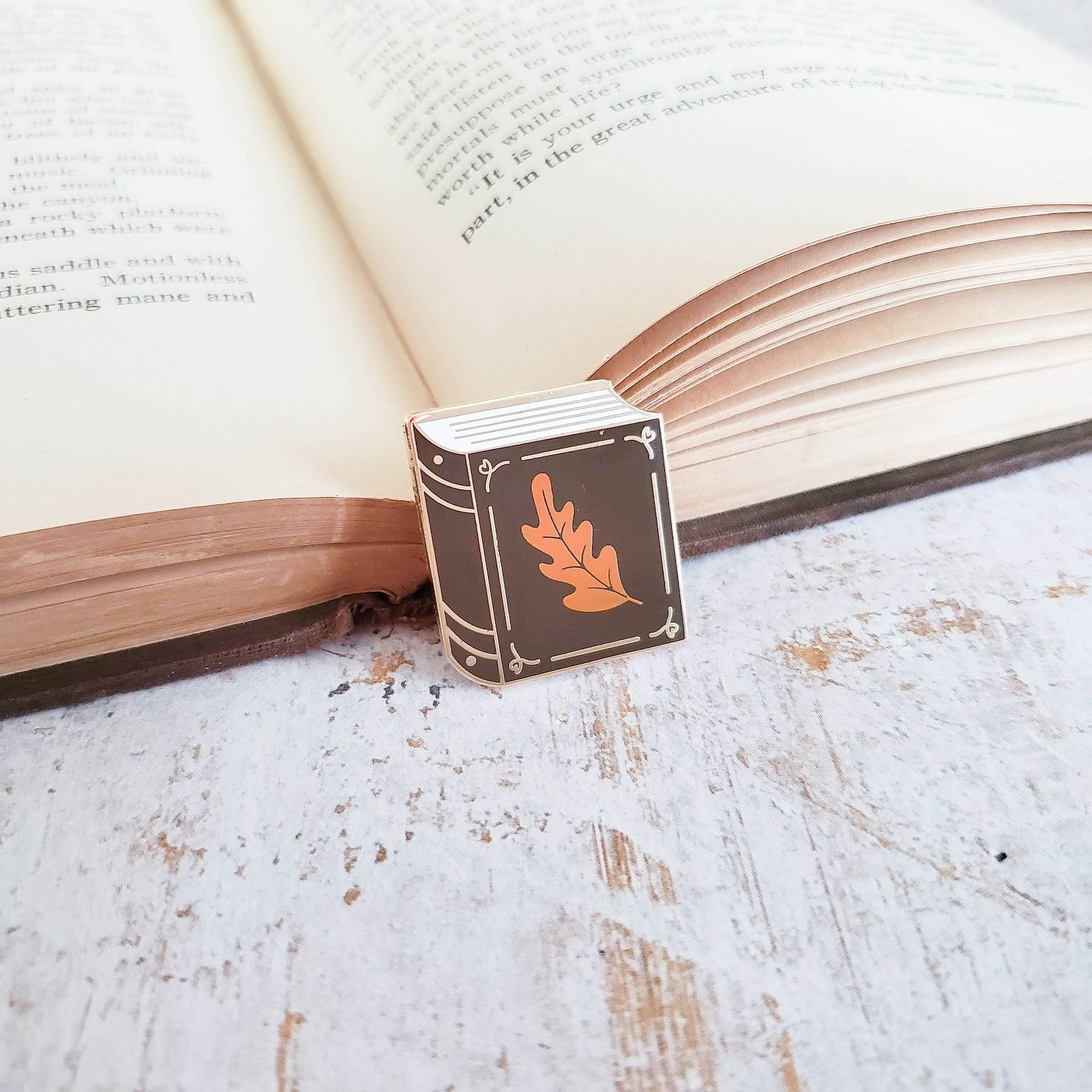 Enamel pin in the shape of a brown book with an orange leaf on it