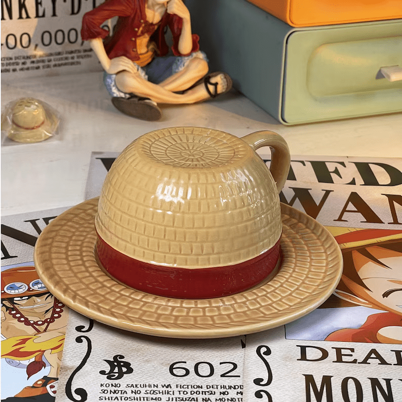 Photo of a mug surrounded by pieces of One Piece merch. The mug has a saucer and the mug placed upside down on the sauce makes it look like a straw hat with a red ribbon.
