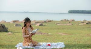 a fair-skinned Asian woman reading on a blanket near a body of water