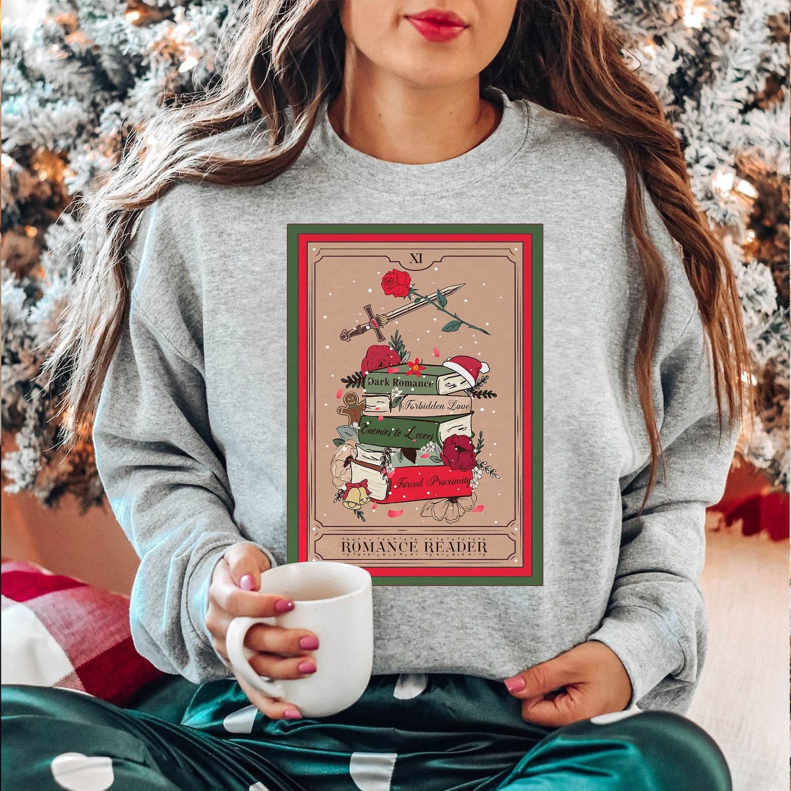 a gray sweatshirt with a tarot card that reads "romance reader" and a stack of books with various tropes on their spines, covered in in snow and with various holiday symbols