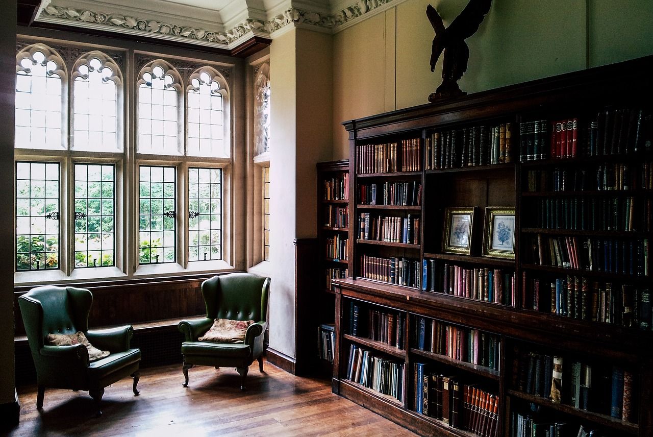 An old-fashioned library in a stately home. On the left side of the picture is a set of eight windows showing a garden, with two green leather chairs in front of them. On the right side is a dark wood bookshelf, filled with books, with some pictures on the shelves and a statue on top.