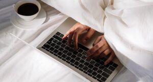 hands reaching out from under a comforter to type on a Macbook. There is a cup of coffee on the bed as well