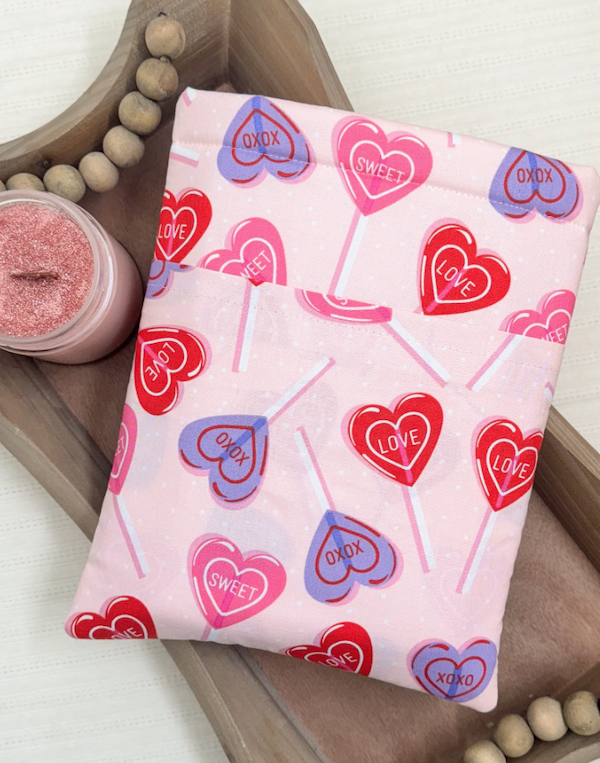 A pink book sleeve with a pattern of red, purple, and pink lollipops that say things like "LOVE," "SWEET," and "XOXO."