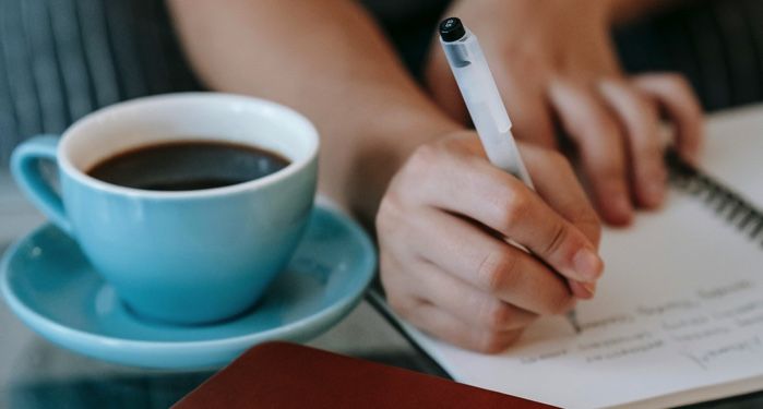 a tan-skinned hand writing in a notebook with a cup of coffee next to them