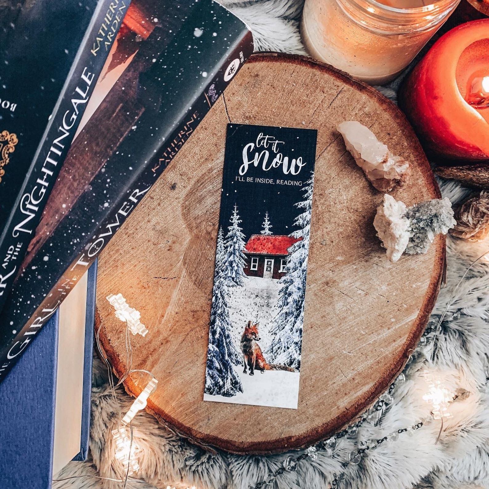 A dark blue bookmark with snow covered trees and a cabin that reads "Let it Snow, I'll Be Inside Reading"