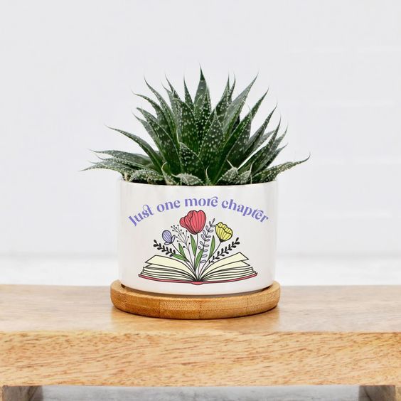 a white ceramic planter with the words "just one more chapter" and an open book with flowers growing out of the pages 