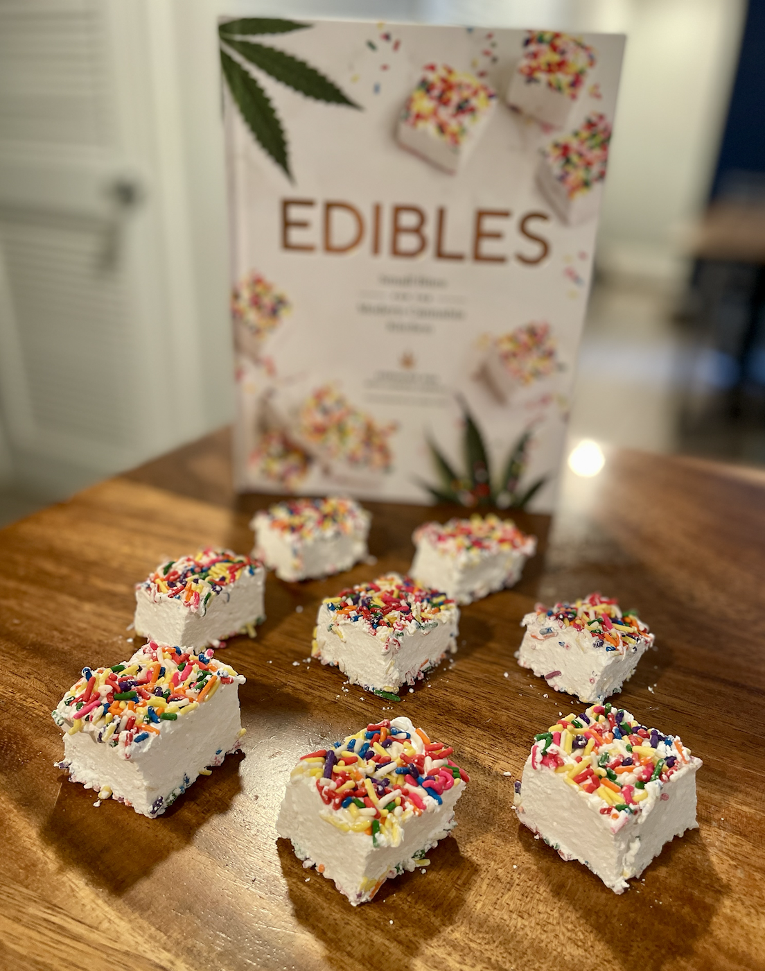 Photo of square homemade marshmallows topped with rainbow sprinkles on a wooden table next to the cookbook Edibles