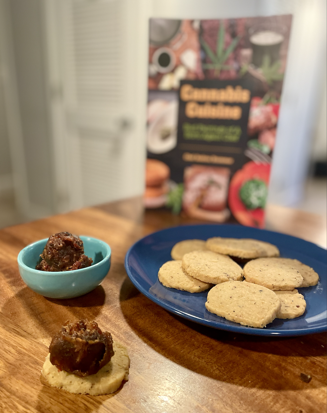 Photo of a small blue plate of homemade crackers, a small bowl of dark bacon jam, and one cracker topped with jam on a wooden table next to the cookbook Cannabis Cuisine
