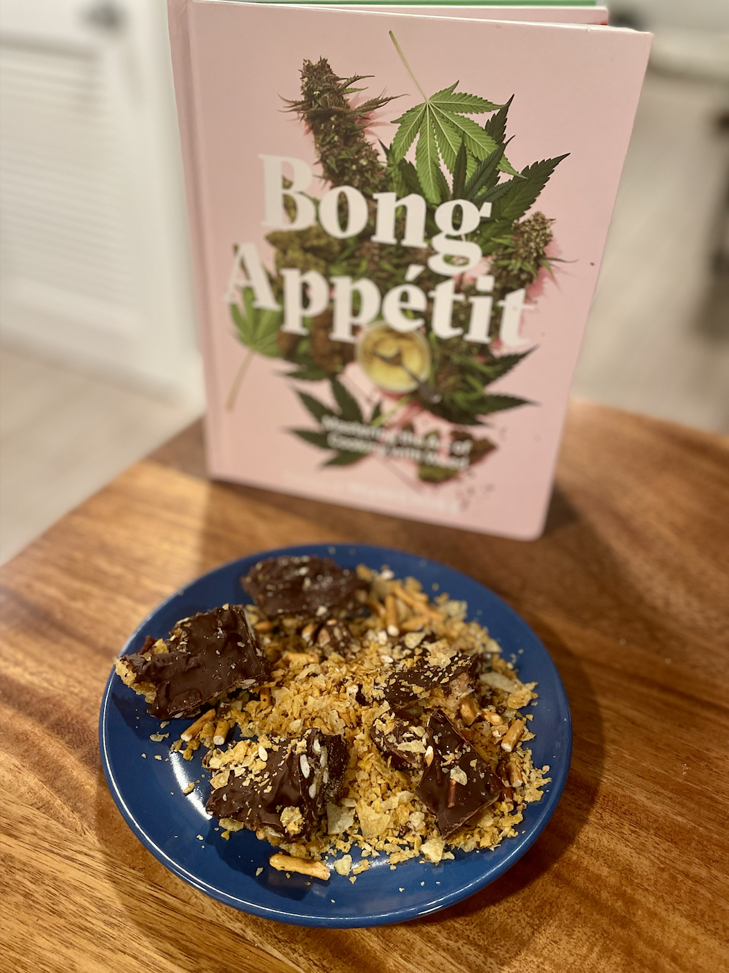Photo of a blue plate of chip and pretzel crumbs and squares of chocolate on a wooden table with the cookbook Bong Appetit