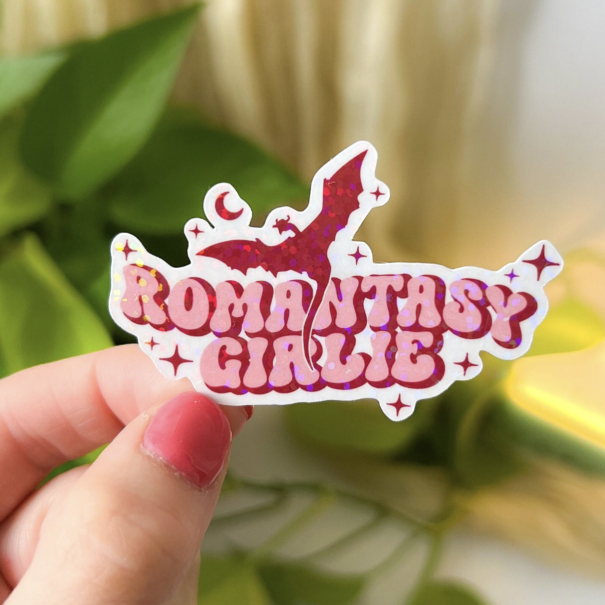 Image of a pink sticker with a dragon that says "romantasy girlie" held up by a white hand with pink nails
