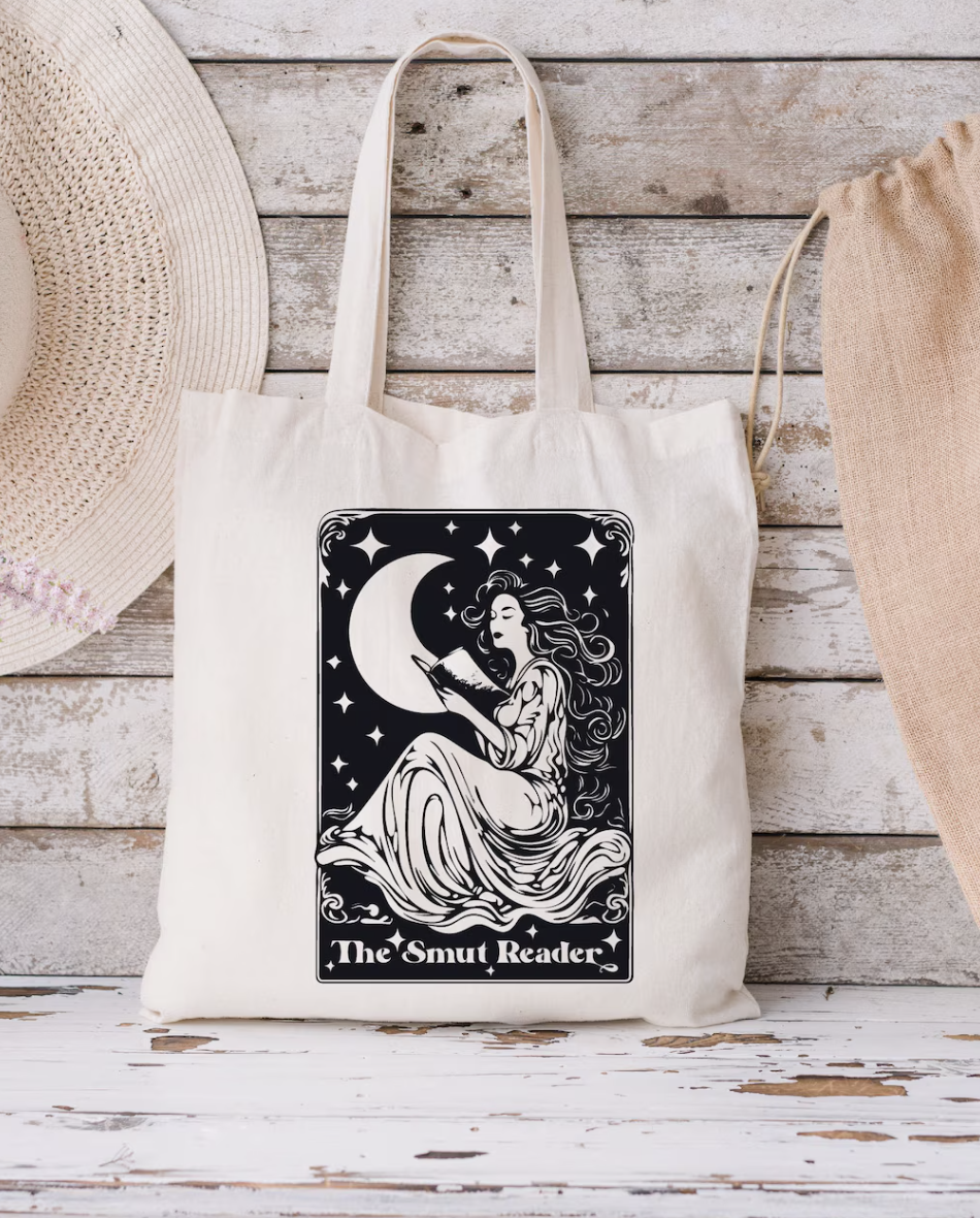 image of a tan tote bag with a black tarot card inspired design of a woman reading a book in front of a crescent moon with the phrase "the smut reader" displayed on a wood plank wall
