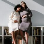 A black woman and an asian woman, boh pregnant, sit close on a bookshelf