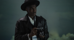photo of a Black man holding an analog camera and wearing a press pass against a foggy background