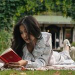light-skinned Asian woman laying on a blanket on the grass and reading