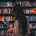 tan-skinned person with long black straight hair reading a book in front of a book store's shelves