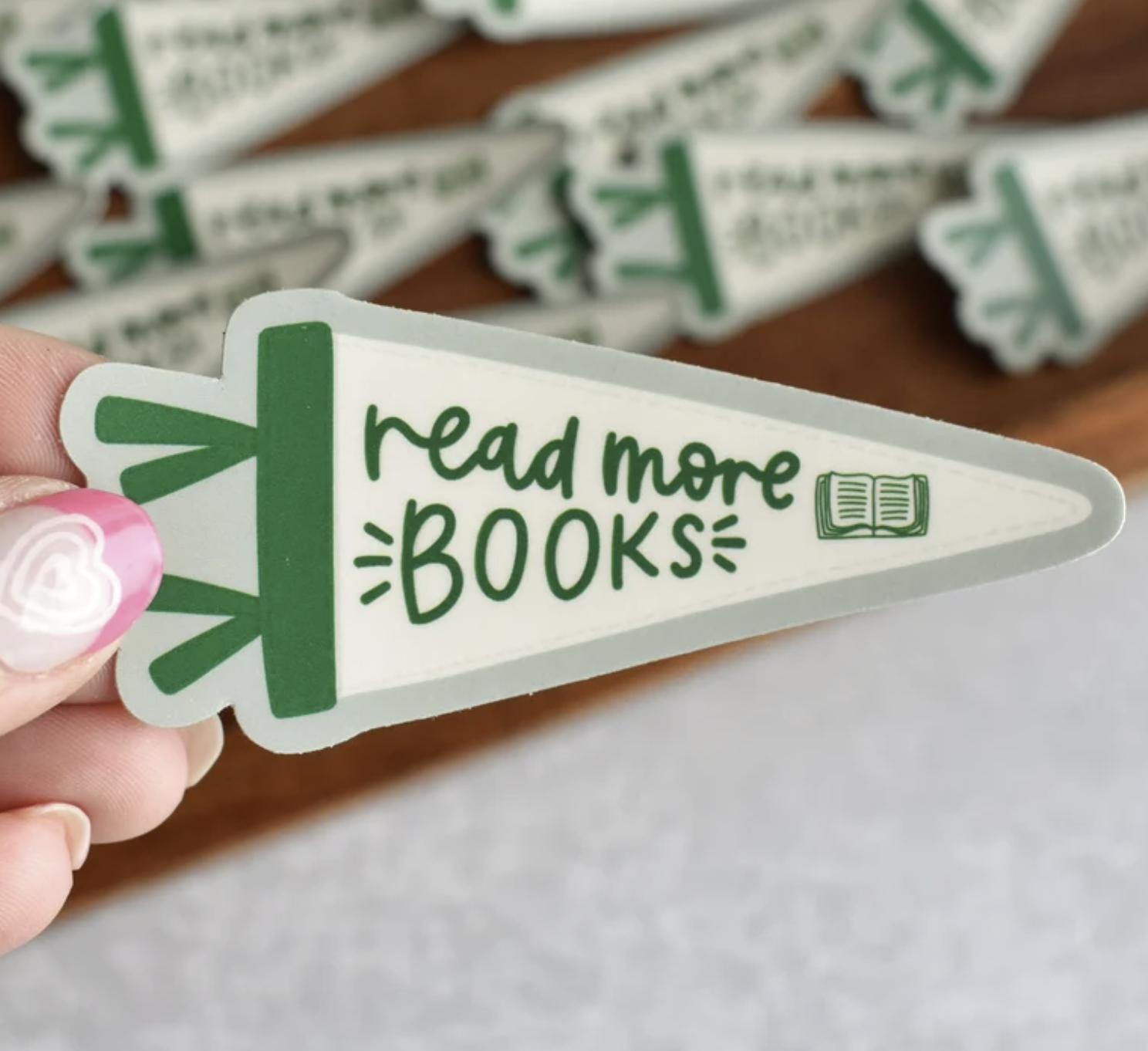 Picture of a white woman's hand holding a sticker in the shape of a pennant with a green stripe that says "read more books"