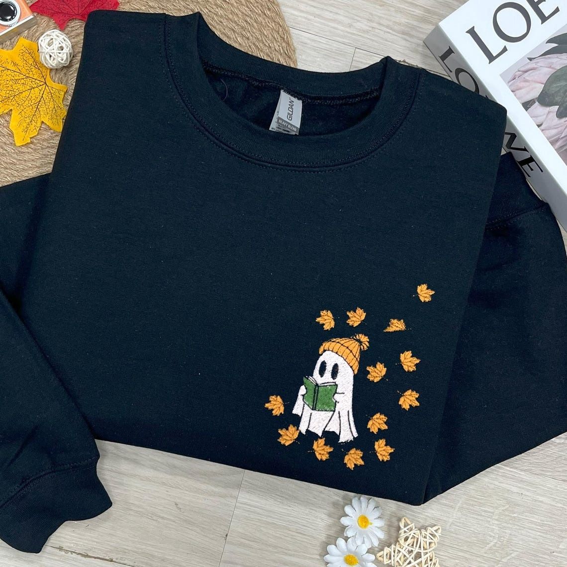 a black sweatshirt with an embroidered reading ghost wearing a beanie and surrounded by falling leaves